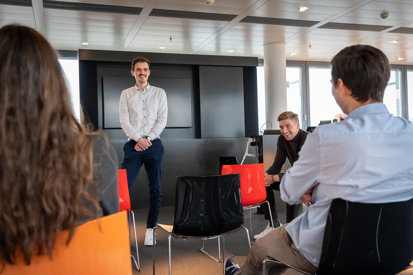 Eine Gruppe von Menschen sitzt auf Stühlen, während ein Mann vor einem Bildschirm steht.