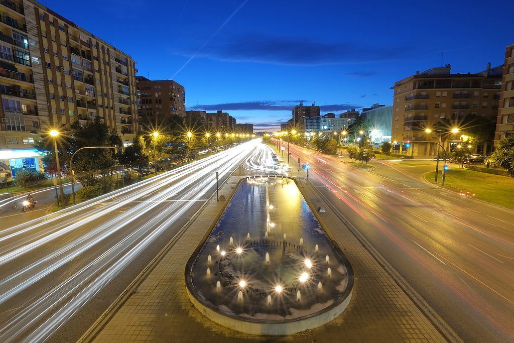 Straße mit einem beleuchteten Brunnen und Straßenlaternen bei Nacht.